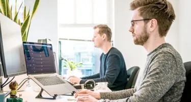 Two designers working side by side at laptops in a bright studio.