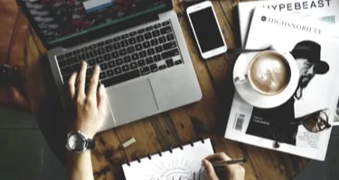 A desk from above with a laptop, a coffee and a designer lettering by hand in a sketchbook.